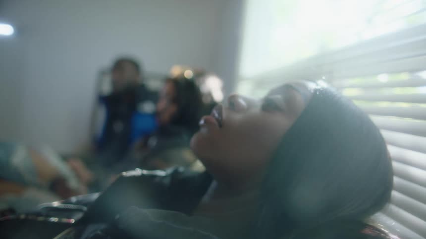A woman sits reclined in a chair in a room with two other people talking in the background and the woman eventually looks right into the camera