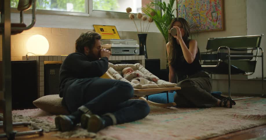 Newborn lies in crib between father with beard turtleneck sweater jeans and shoes and mother with brown hair black top and brown pants drinking from cup sitting on rug in the living room at home