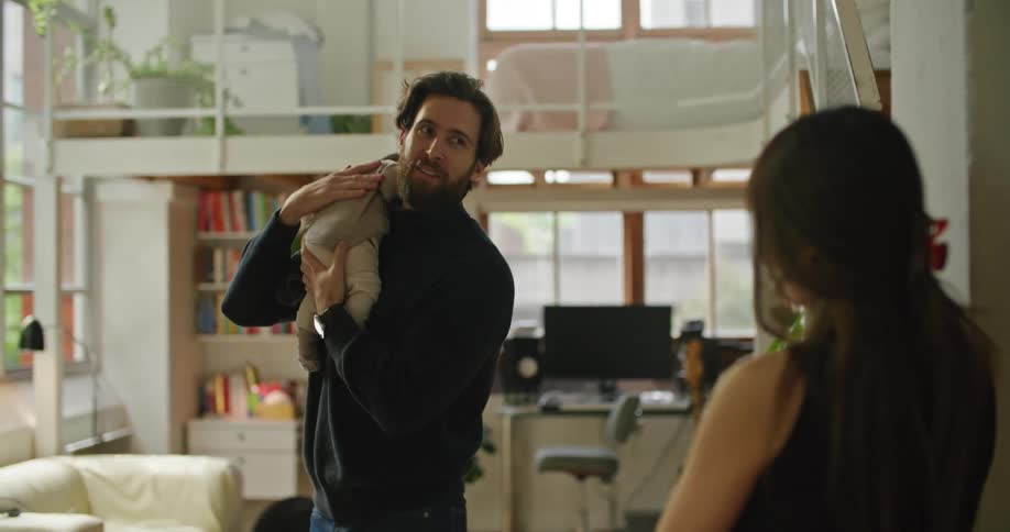 A man burps his baby while standing in the kitchen with his wife