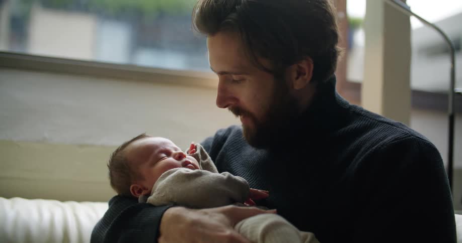 A man sits on the couch while rocking an infant baby in his arms and looking at him lovingly