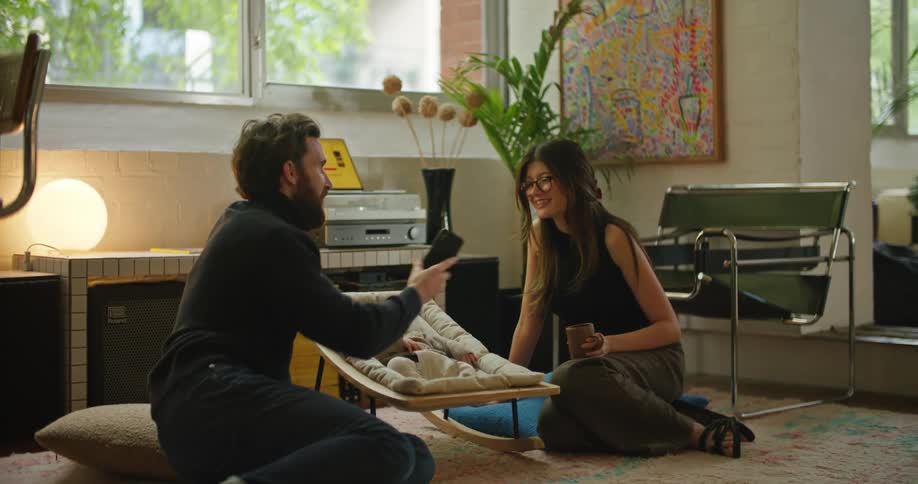 Young bearded man shows something on his smartphone to his wife while taking care of the baby at home