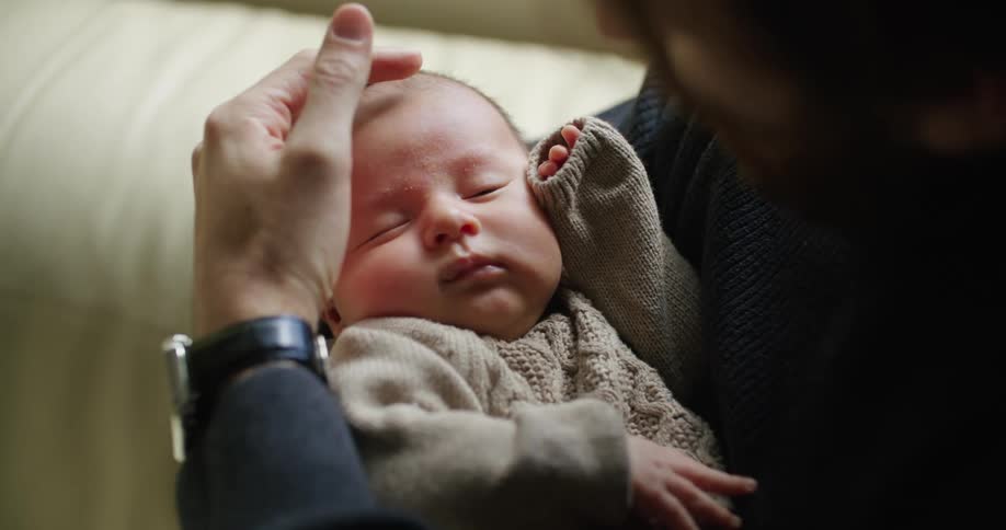 Father sitting on the couch at home rubs the forehead of his newborn baby