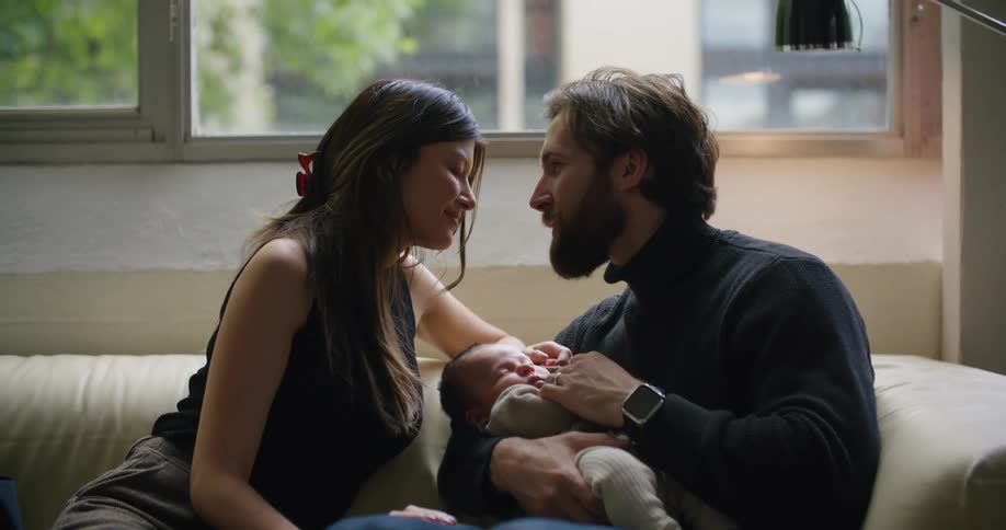 A man cradling a baby while sitting on a sofa leans forward to kiss the woman sitting beside him