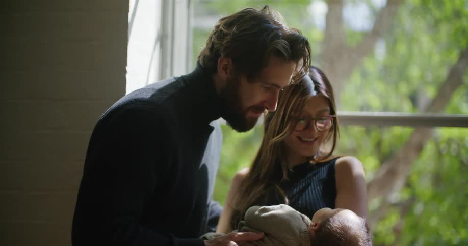Young bearded man to her wife holding their newborn baby at home near a window