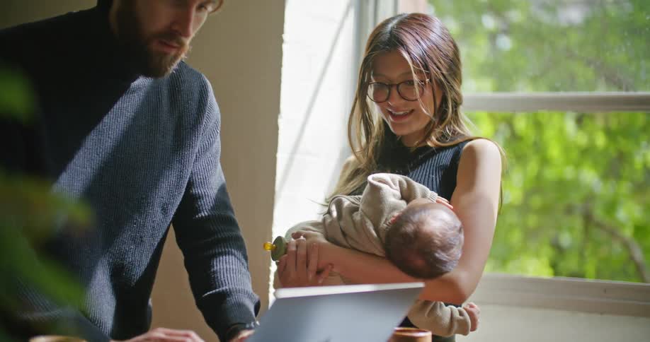 Young bearded man searching on a laptop next to her wife holding their newborn baby at home