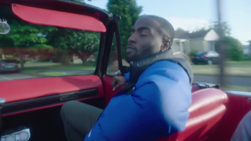 A man is sitting in the passenger side of a red convertible car as it travels through a neighborhood