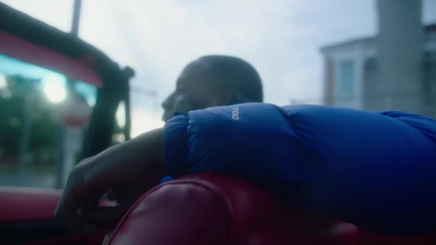 A man is sitting in the passenger side of a red convertible car as it travels through a neighborhood