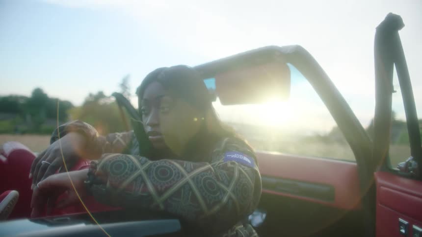 A woman sits facing backward in a convertible car parked in a field during the day