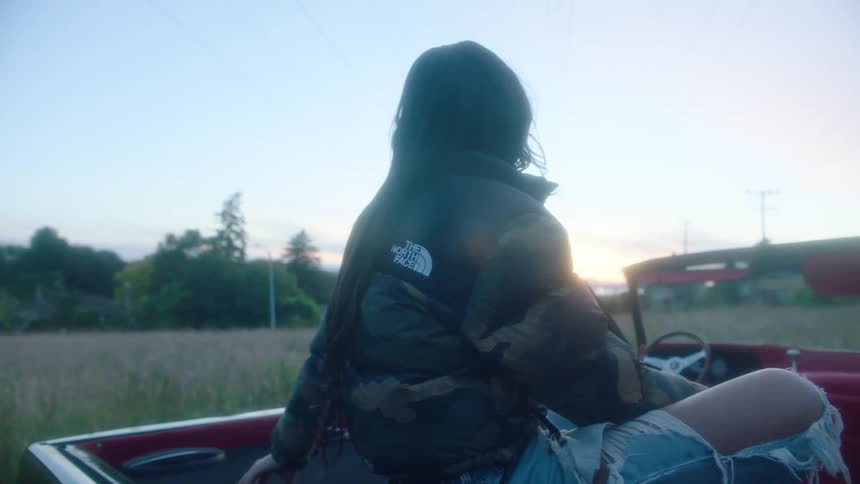 Woman with green coat and ripped jeans sits in convertible car with the roof down in the field at sunset