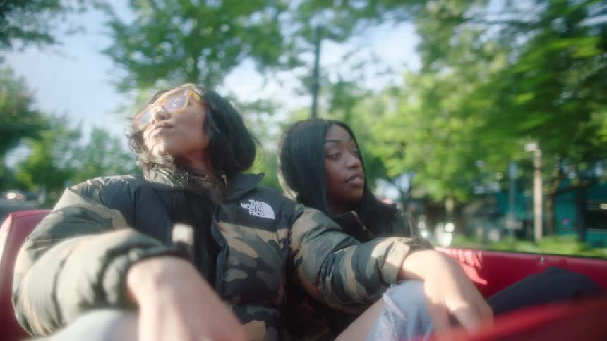 Two women ride in convertible passing by small shops