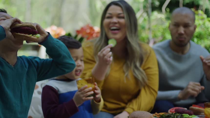 Family eating burgers and fresh vegetables sitting on a couch in the backyard at daytime