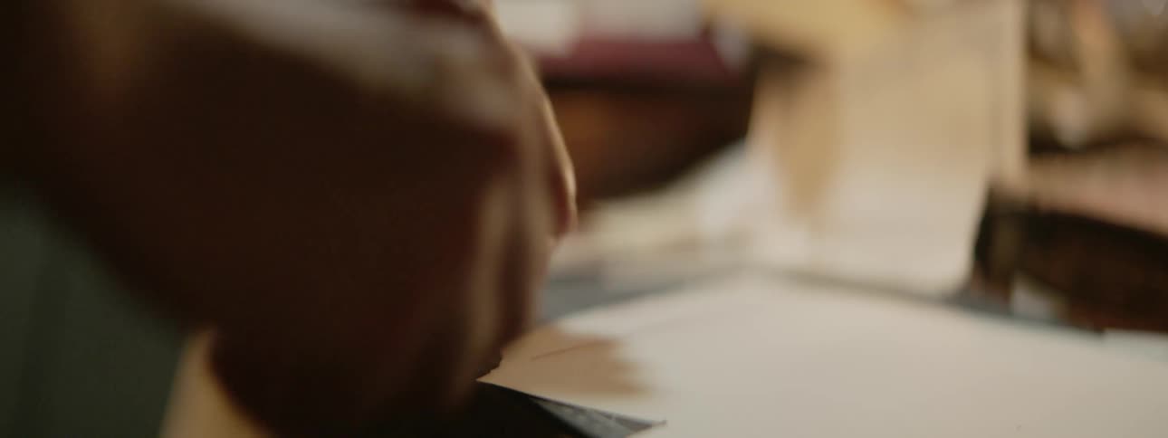 A man is sitting at a table as he picks up a pair of scissors and cuts paper