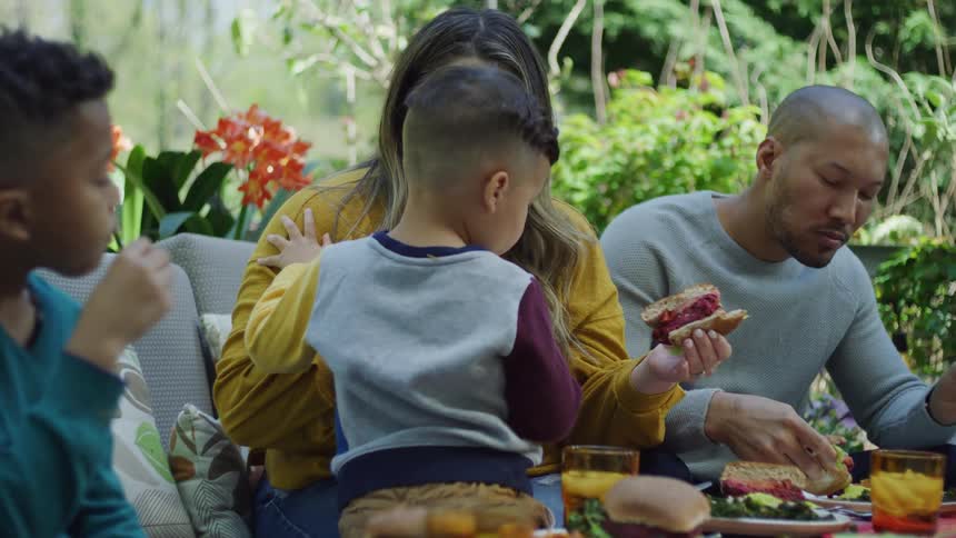 Family sitting on a couch in the backyard eating hamburgers at daytime