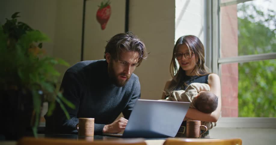 Young bearded man searching on a laptop next to her wife holding their newborn baby at home