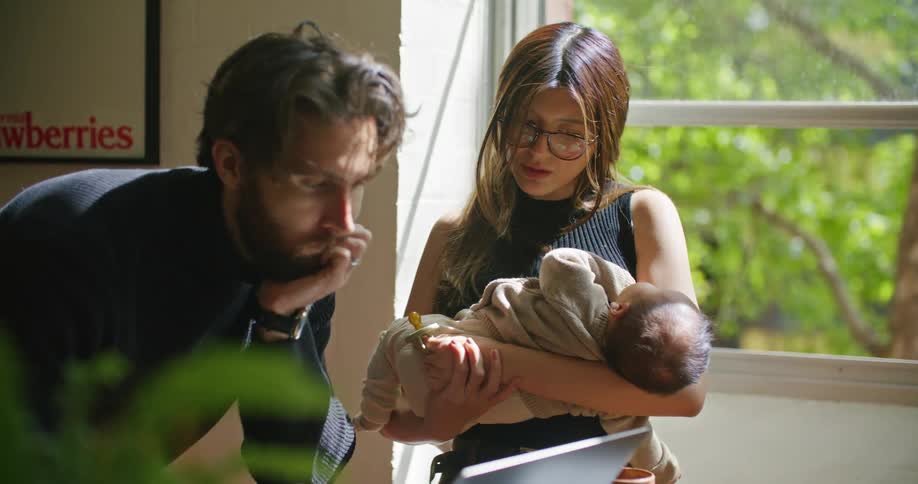 A man uses a laptop indoors while a woman rocks a baby in her arms next to him