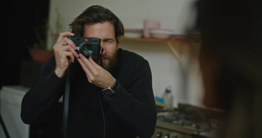 A man smiles while taking a photo of his partner in the kitchen