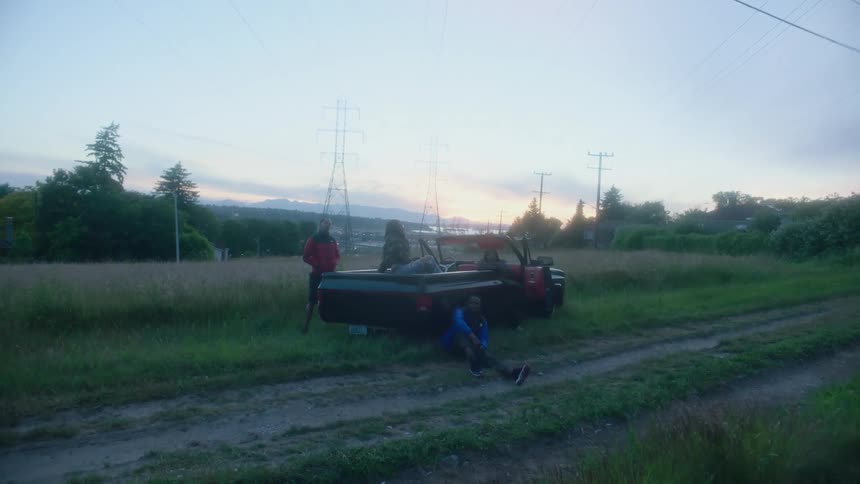 Friends sit and stand around convertible parked near dirt road with red sky in the background