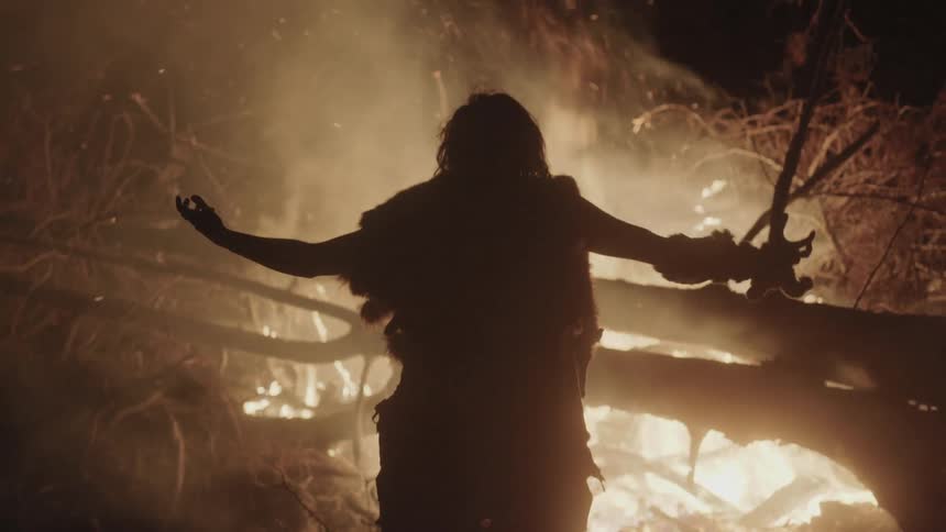 Wilderness man raising his arms while standing in front of a fire at night