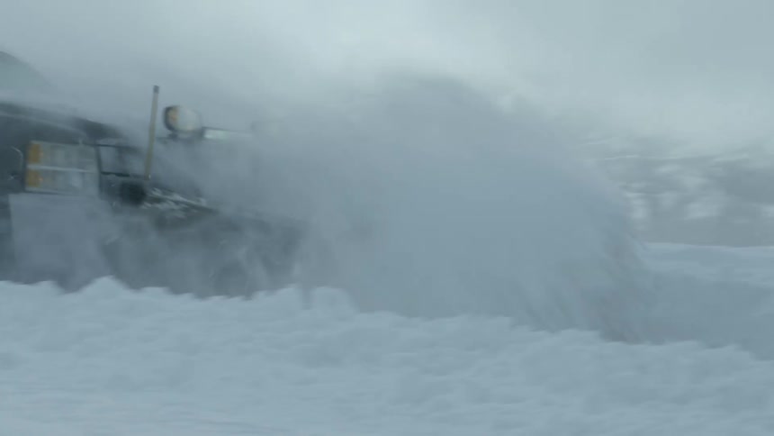 Snow flies into the air as a truck with a snow plow travels through a road with mountains in the distance
