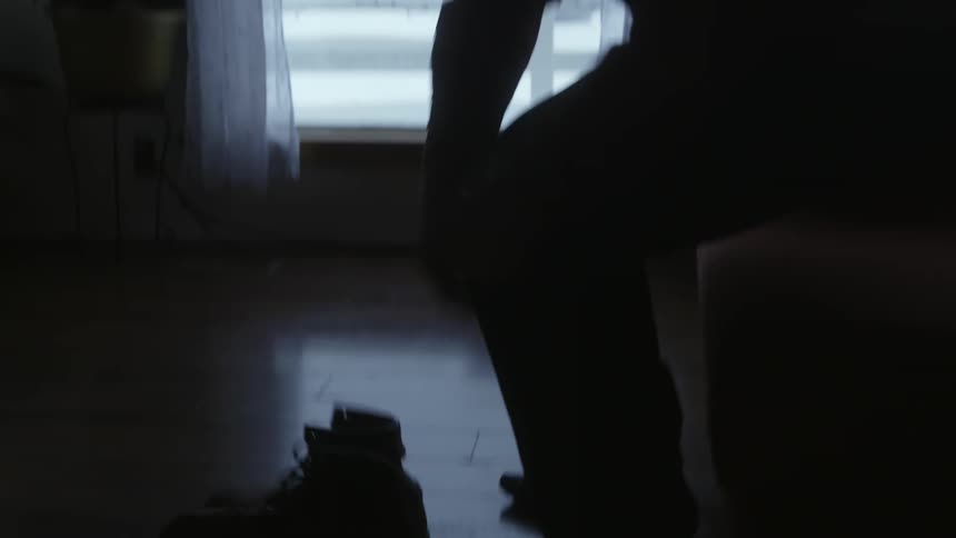 A man at home sets his boots on the floor and sits down to put them on