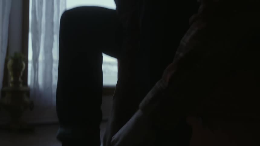 A man is sitting down putting on his boots as light shines in from the window