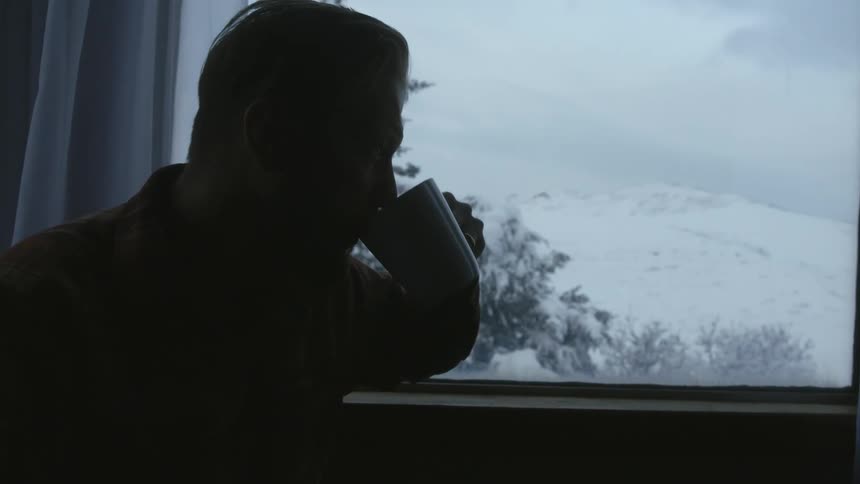 A man is sitting down drinking a cup of coffee as he looks out the window at the snow covered trees and hill