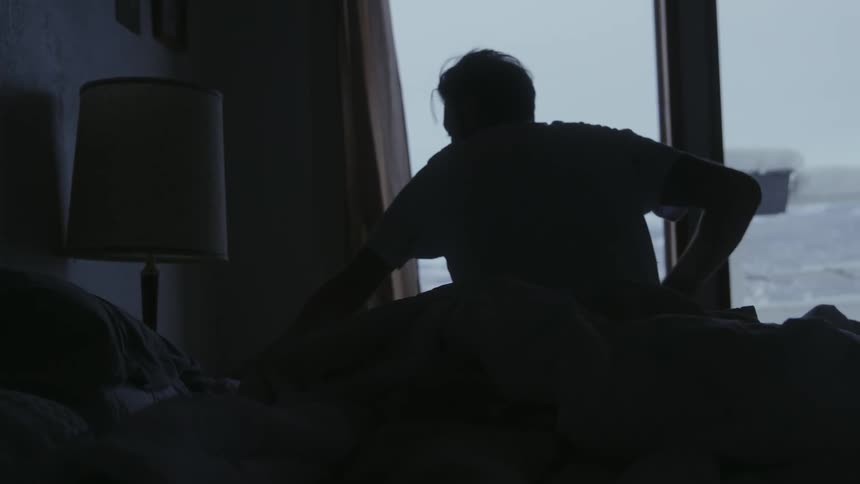 A man gets up out of bed and sits on the side of his bed looking out the window at the snow outside