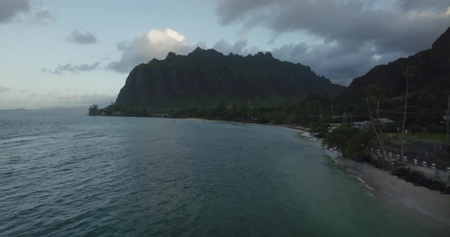 People are on the beach and traffic moves on the street along the coast as waves roll in and there are mountains along the shoreline