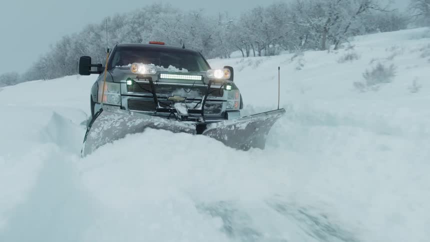 A truck has a plow attached to the front and is pushing deep snow off the roadway during a winter storm