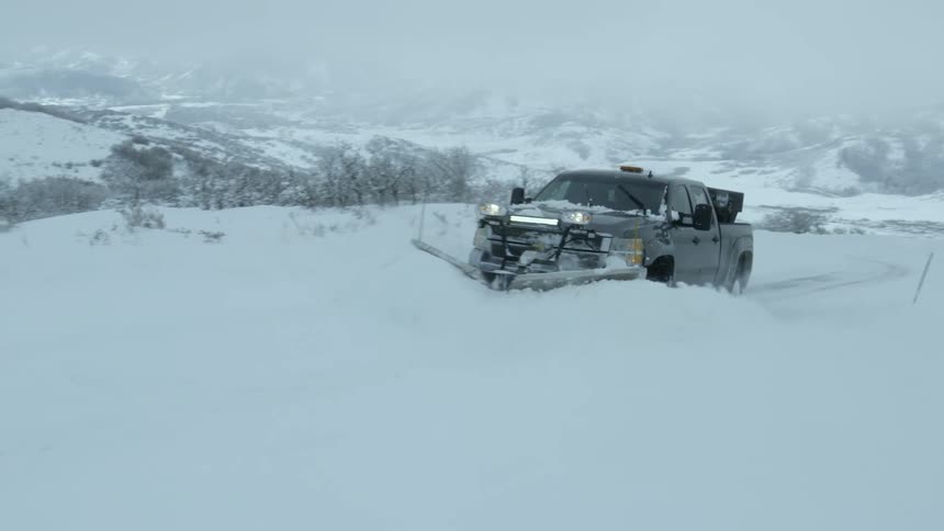 A truck with a snow plow affixed to the front clears deep snow from a countryside road on a foggy winter day