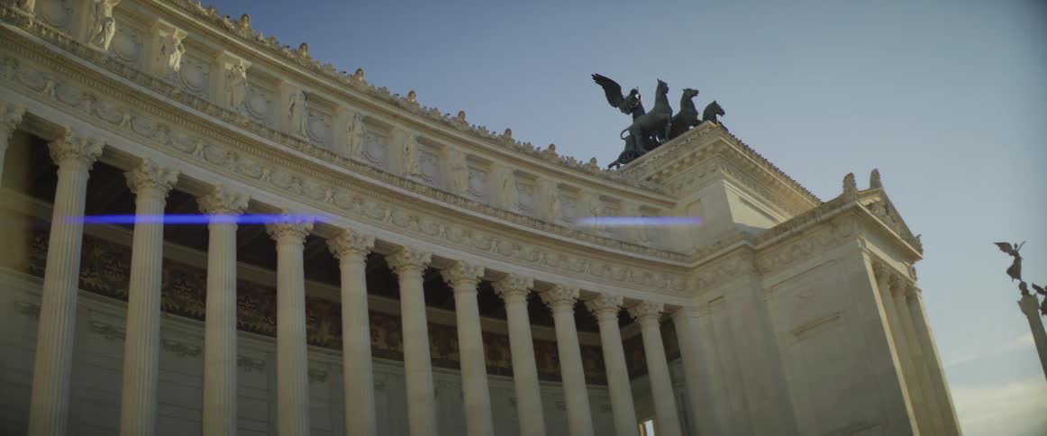 Large white building with classical architecture topped with statues stands near columns also topped with statues under a sunny sky