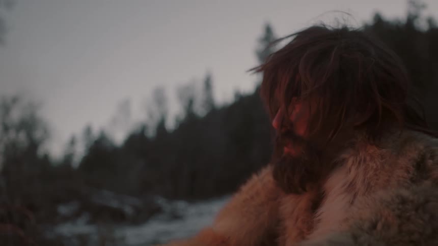 A man wearing fur sits beside a fire