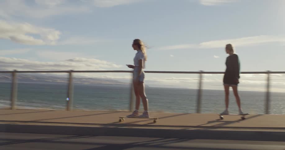 Two teenage girls are riding skateboards on a road overlooking the ocean
