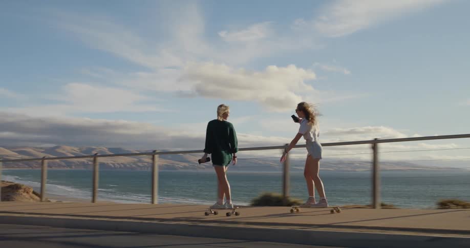 Two young teenager girls wearing shorts ride skateboards down the hill next to the ocean