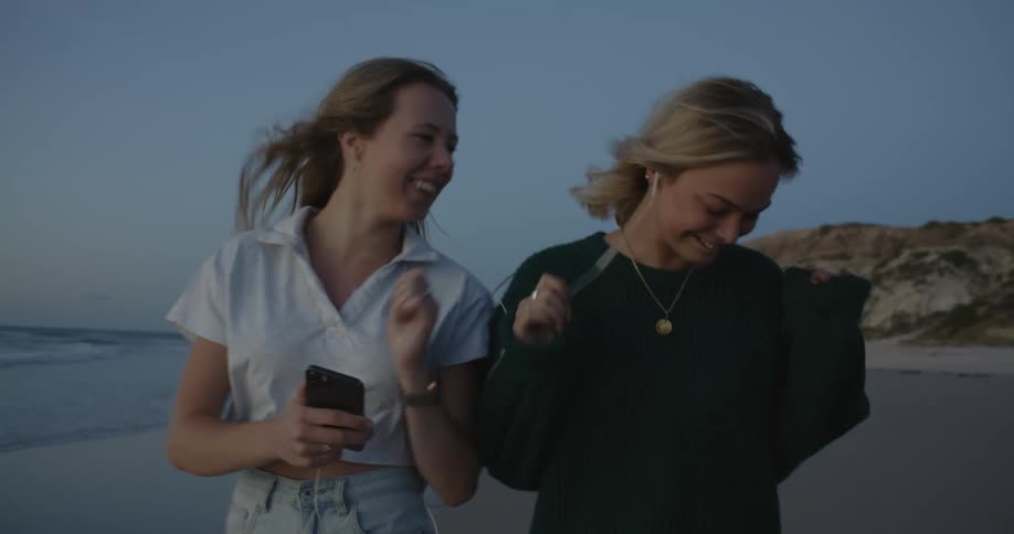 Two female friends share earbuds while walking along beach