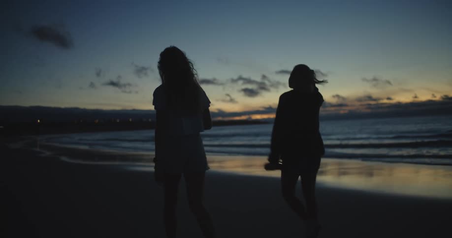 Silhouette of two young women walking on a beach next to the ocean at sunset