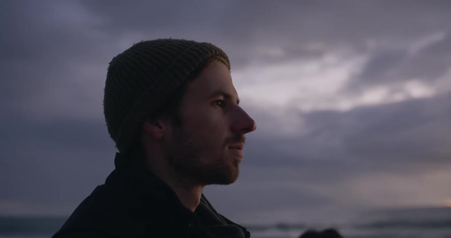 Close up of the face of a young man standing on the beach looking at the horizon at evening