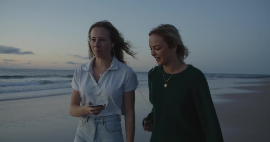 Two young happy friends laugh and talk looking at a phone while walking on a beach at evening