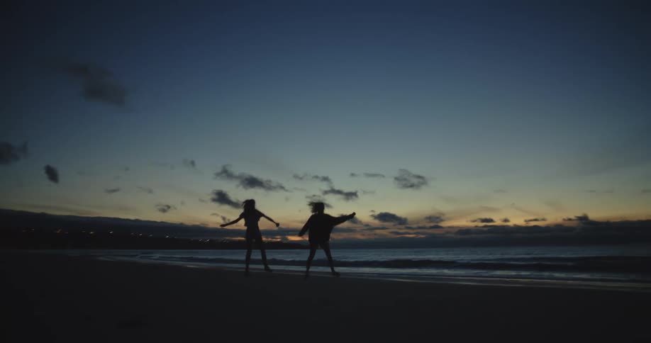 Two young women dance and jump on the beach next to the ocean at sunset
