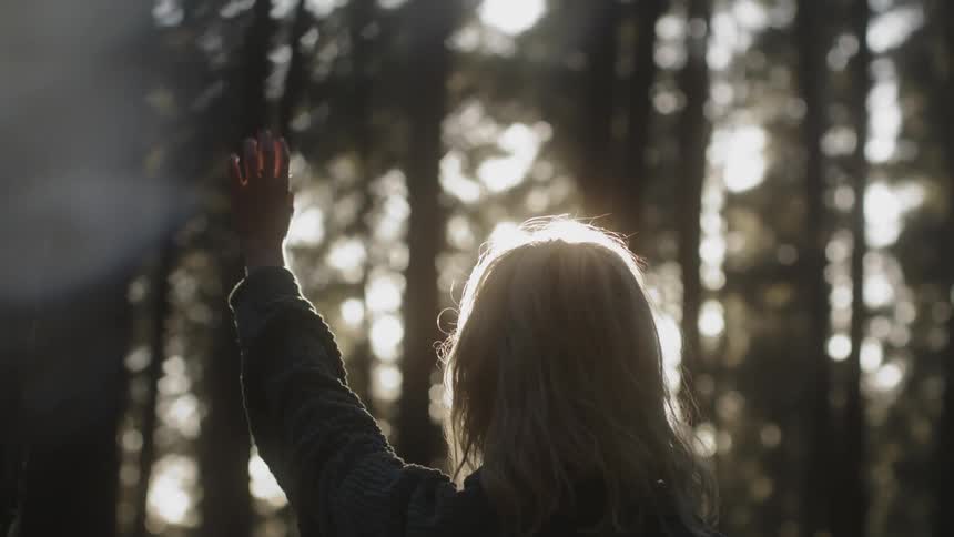 A teenage girl is standing in the woods holding up her hand as the sun shines brightly through the trees
