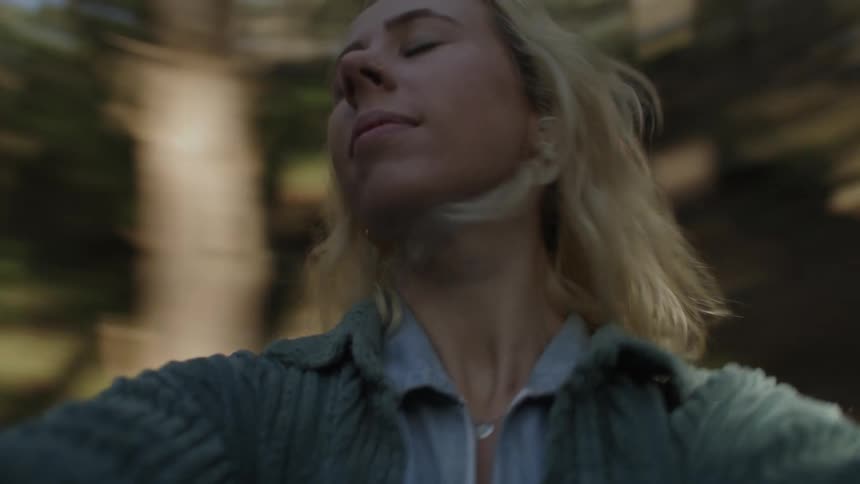 A blonde woman with head back and eyes closed spinning around in the woods