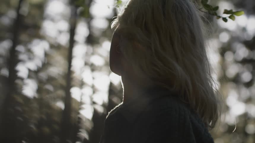 A teenage girl is standing in a forest and her hair moves in the breeze as the sun shines on the trees