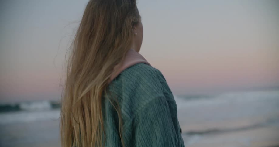 A woman with long blond hair is walking along the beach as the waves roll into shore