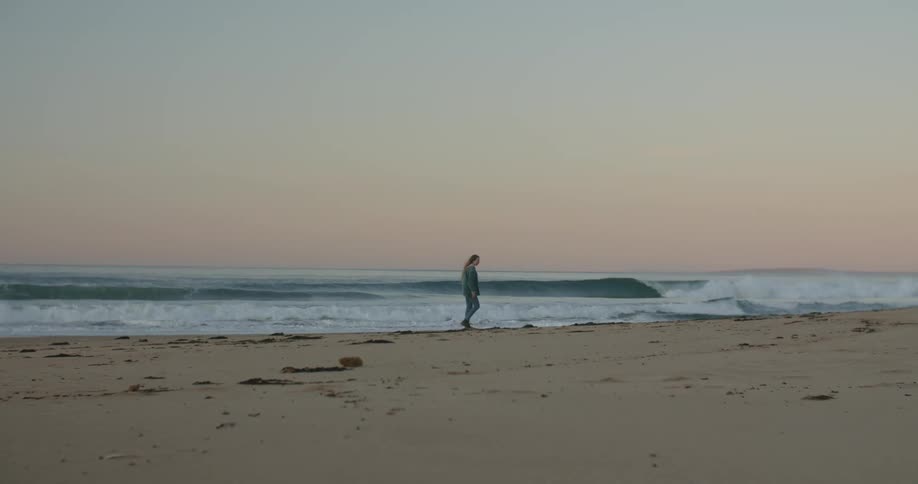 Woman with brown hair denim jacket and jeans walks along beach near the wavy ocean at dusk