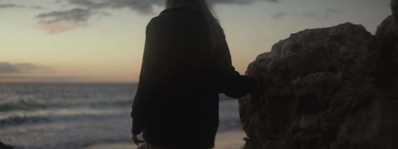 A woman navigates large rocks and boulders on a beach at sunset