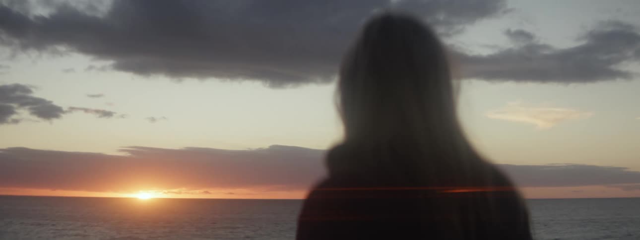 A woman is standing on the beach looking at the sun on the horizon