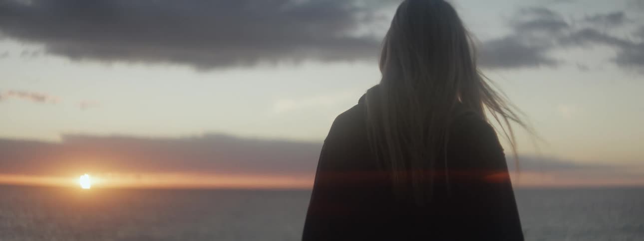 A teenage girl is standing on the beach looking at the sun on the horizon