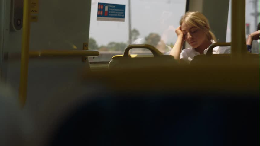 A teenage girl is sitting on a train and she rubs her eyes as she leans her head against the window