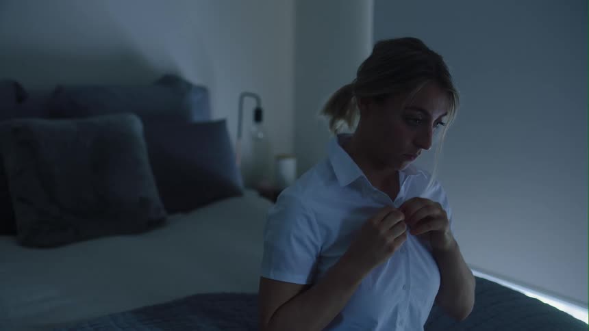 A woman sits on a bed and buttons her shirt