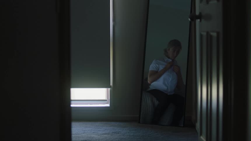 Woman with gray hair and jeans buttons her blue blouse in dark room